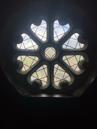 A low-angle shot depicts a circular rose window set into a dark, possibly stone, structure. The window itself consists of a central circular pane surrounded by seven outward-facing, stylized heart shapes. Each pane is divided by dark, thin lines into geometric patterns, primarily triangles and squares. The window panes transmit light from the outside, revealing a mix of sky and vegetation. The surrounding structure is largely obscured by shadow, with only segments of the window frame and adjacent wall visible. The scene takes place in Bradford, United Kingdom.