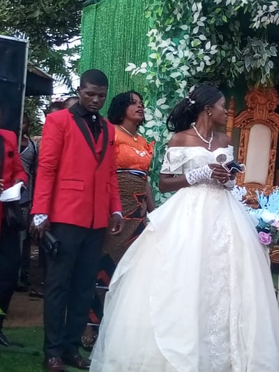 This image captures an outdoor event in Amokvo, Nigeria, featuring three primary individuals positioned against a decorated green backdrop.
On the left, a dark-skinned male, with short dark hair, stands wearing a red blazer with black lapels, a black waistcoat, a black shirt with a silver bolo tie, and black trousers. His right hand holds a black clutch bag, and a white patterned cuff is visible on his wrist. His gaze is directed downwards.
Adjacent to him, a dark-skinned female with curly dark hair wears an orange lace top with ruffled shoulders and a multi-patterned skirt. She has a gold necklace and appears to be speaking or singing, with her mouth slightly open and her head tilted upwards and to the right.
To the right, a dark-skinned female, with dark hair styled in a ponytail, is dressed in a white off-the-shoulder ball gown with intricate lace detailing. She wears a pearl necklace and white lace fingerless gloves. In her left hand, she holds a white, circular object and a black face mask. Her head is turned to the right, away from the direct view of the camera.
The background comprises a green, vertically-striped fabric backdrop ornamented with artificial green foliage. To the far right, a portion of an ornate, carved wooden chair is visible, and a floral arrangement with blue and purple elements is situated nearby. A black speaker unit is partially visible on the far left. Natural tree foliage is present above the constructed backdrop, and the ground in the foreground is covered in green, resembling grass.