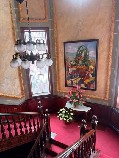 This is a high-angle interior shot of a grand staircase in La Orotava, Spain, likely within a historic building. The staircase features dark wooden banisters with intricately carved spindles and is covered in a vibrant red carpet. The walls are paneled with dark wood at the lower level, transitioning to a decorative marble-effect finish with ornate gold frames at the upper levels. A large framed painting depicting a festive scene with people and oxen is prominently displayed on the wall. Above the staircase, a multi-light chandelier with frosted globes hangs from the ceiling. A tall window with a grid pattern is visible behind the chandelier. On a marble-topped table near the bottom of the stairs, a vase filled with pink anthuriums and other greenery is present, with another potted plant of white anthuriums beside it. The scene appears to be well-maintained and opulent.