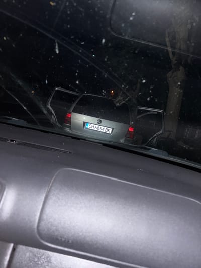 The image shows the rear of a silver Volkswagen station wagon with its rear doors open, parked at night on a street in Blatets, Bulgaria. The car's license plate reads "CH 6864 BK". The scene is viewed from inside another vehicle, looking through the windshield, which is speckled with water droplets or dirt. A tree with visible branches is to the right of the car. The foreground shows the dashboard of the vehicle from which the photo was taken.