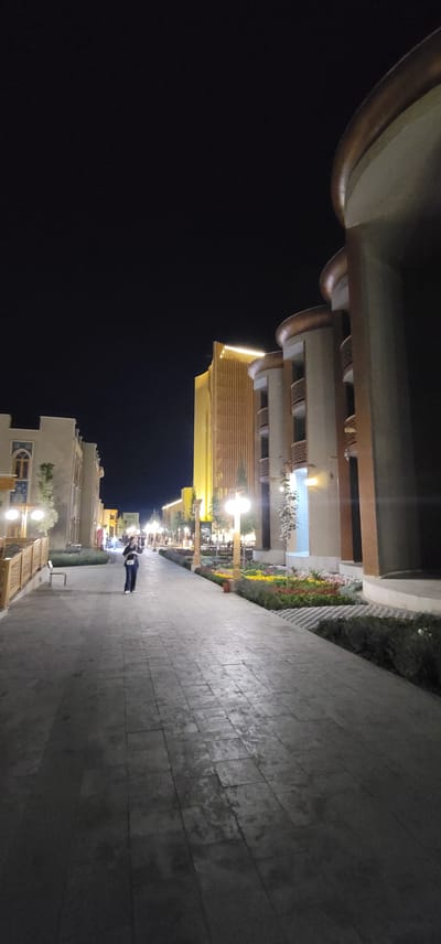 The image shows a pedestrian walkway at night in Satkak, Uzbekistan. The walkway is paved with large, rectangular tiles and is lined with trees and flower beds. On the right side of the walkway, there are several modern buildings with curved balconies and decorative elements. The building in the center is tall and illuminated with warm, vertical lighting. On the left side of the walkway, there is a lower building with arched windows. A single person is walking away from the camera in the middle of the walkway. Streetlights illuminate the scene, casting a warm glow on the surroundings. The sky is dark and clear.