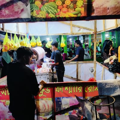 This image depicts a vibrant night market scene in Charfasson, Bangladesh, centered around a food or refreshment stall. In the foreground, two young male vendors in black t-shirts are engaged in activities behind a counter. The vendor on the left is seen from the back, while the one in the middle, wearing clear gloves, appears to be preparing an item. A prominent feature of their stall is a large, rotating cylindrical machine, likely used for making cotton candy, with pink and white material visible inside.

Above the stalls, numerous green mesh bags are suspended, filled with various fruits such as bright yellow/orange citrus and lighter red/yellow fruits. The upper part of the image shows colorful banners; one prominently displays an array of fresh fruits including watermelon, grapes, oranges, and pineapple, while another shows a pink, frothy dessert. A red banner below, visible across the stall, features yellow Bengali text that partially reads "খাদ্য কাশ্মীরি রোন" (Khaddo Kashmiri Ron), suggesting a type of food or brand.

In the middle ground, an older man in a blue polo shirt stands behind an adjacent section of the market stall. Further back on the right, a woman wearing a black hijab and a patterned dress is seated with a child, possibly customers. The setting is illuminated by artificial lights, indicating that the market operates in the evening or at night. In the background, green pillars with the partial word "CLEMON" (likely a brand name) are visible. The overall scene conveys a lively and bustling atmosphere common to a local fair or market.