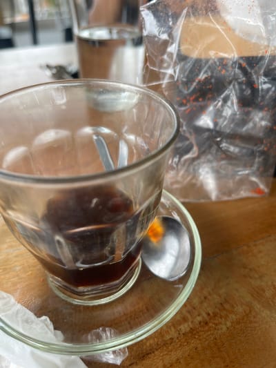 The image shows a close-up of a glass of coffee on a saucer, placed on a wooden table. A plastic bag containing a reddish-orange powder is visible in the background. A spoon rests on the saucer. A glass of water is also visible in the background, slightly out of focus. The scene appears to be indoors, possibly a cafe or restaurant. There is no indication of the city of Pulokiton, Indonesia, or any specific details about the environment that would place it there.