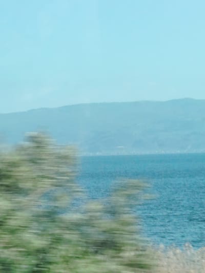 This media file captures a serene landscape viewed from a moving perspective, characterized by pronounced motion blur in the foreground.

The scene is composed of three main layers: a blurred foreground, a clear body of water in the midground, and distant mountains under a light blue sky. In the immediate foreground, horizontal streaks of green and brown indicate rapidly passing vegetation or ground cover, a clear sign that the photograph was taken from a vehicle in motion.

Beyond the blurred foreground, a calm body of water, likely a large lake, stretches across the midground. Its surface appears in varying shades of blue and turquoise, with subtle textures suggesting gentle ripples or small waves. In the background, a range of hazy, bluish mountains or hills extends across the horizon, softened by atmospheric perspective. A faint, light-colored line along the base of these distant hills might suggest a far-off settlement or structures on the opposite shore. The sky above is a uniform, clear light blue, indicating bright daylight.

The image conveys a sense of peaceful travel through a natural setting. The motion blur contrasts with the tranquility of the distant landscape. There is no visible text in the image. Given the location context of Iznik, Turkey, this is likely a view of Lake Iznik and its surrounding topography.