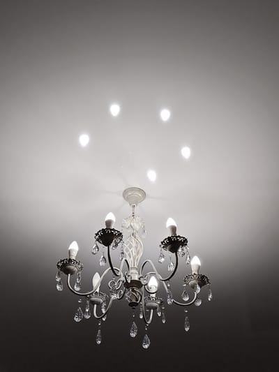 The image shows a chandelier hanging from a white ceiling. The chandelier is ornate, with multiple arms and decorative crystal droplets. There are six lights on the chandelier, all illuminated, and their reflections are visible as bright spots on the ceiling. The lower portion of the image is dark, suggesting the presence of a room below the chandelier. The exact location in Balqıq bï, Kazakhstan cannot be determined from the image alone.