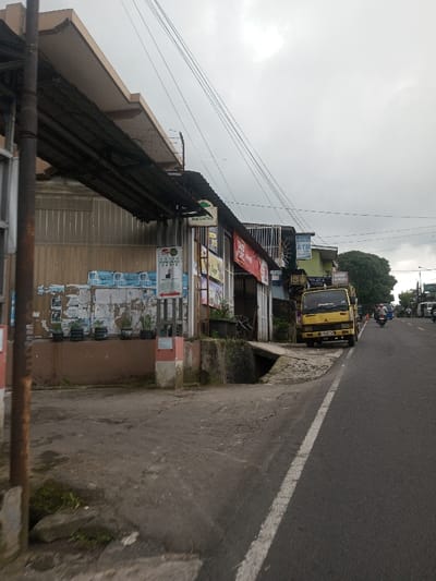 A street scene shows a yellow truck parked beside a road. A storefront lined with signs and advertising sits on the left, displaying posters and a pile of what appear to be water bottles. An awning overhangs the building. Power lines stretch across the overcast sky. Other vehicles and pedestrians are visible further down the road. The road itself has a white line marking its edge.