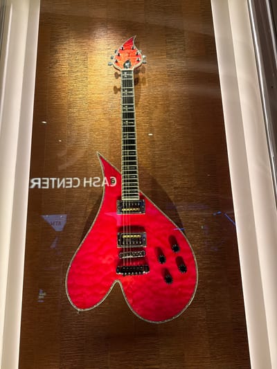 A red heart-shaped electric guitar hangs horizontally against a textured, brown-toned wall. The guitar features a headstock with tuning pegs, a fretboard with inlays, pickups, and black control knobs. The reflection on the wall contains the text "CASH CENTER" reversed. The scene is illuminated by both direct and reflected light. It is located in the city of Hollywood Seminole Reservation, United States of America.