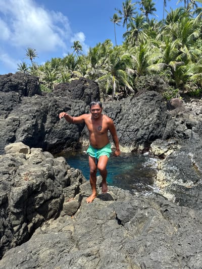 A man in green swim trunks stands on a dark rocky shore, posing for a picture while partially immersed in water. The surrounding landscape consists of rugged, black volcanic rocks, with lush green palm trees visible in the background, suggesting a tropical location. The sky is blue with some scattered clouds. The location is the city of Santa Monica, Philippines.