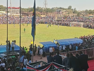 This image depicts a bustling football match taking place in an outdoor stadium in Siaya, Kenya. The stands and surrounding embankments are densely packed with a large crowd of spectators, indicating a significant event.
On the bright green football pitch, players in both green and white/light-colored jerseys are actively engaged in a game, with goalposts visible at either end. Along the sideline, two blue covered dugouts are present, surrounded by a mix of officials, security personnel, and other individuals.
In the immediate foreground, a viewing area features a decorative banner displaying the black, red, and green colors of the Kenyan flag. A tall flagpole in the mid-ground proudly flies a large blue and yellow/gold flag with a distinct central emblem. Numerous media personnel equipped with cameras and tripods are strategically positioned, some atop vehicles, to capture the event. Security personnel are also visible throughout the lower areas of the stadium.
The scene is well-lit by bright sunshine under a clear sky, suggesting daytime, likely late morning or early afternoon. In the background, beyond the stadium, various buildings and structures indicate an urban or semi-urban environment, along with visible utility poles and electricity pylons. The overall impression is one of a vibrant public gathering centered around a sporting event.