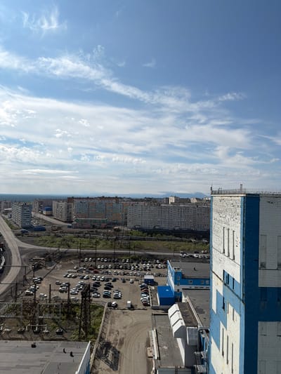 This is an aerial, eye-level, outdoor, daylight, high-angle shot of Norilsk, Russia. The scene captures a section of the city characterized by apartment blocks, industrial infrastructure, and a large parking lot filled with vehicles. The apartment buildings in the midground are multi-story structures with varying facades, indicating a mix of colors and architectural styles. A prominent feature is a large, white and blue industrial building in the foreground, dominating the right side of the frame. Its exterior appears weathered. Surrounding the buildings are roads, power lines, and some vegetation. The sky above is a clear blue with scattered white clouds. The overall impression is of a functional, somewhat stark urban landscape, likely a residential or industrial area within Norilsk. There are no visible people in the frame.