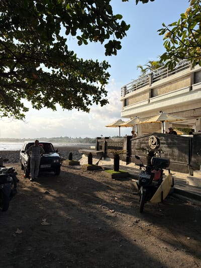 This is a medium shot of a beachfront scene in Pekutatan, Indonesia. In the foreground, a man stands next to a dark-colored SUV parked on a dirt and gravel lot. The man is wearing a plaid shirt and light-colored pants. A motorcycle with a surfboard attached is parked to the right of the SUV. In the background, a modern building with multiple levels and a balcony overlooks the ocean. Several umbrellas are set up on the balcony. The ocean is visible with a cloudy sky above. The ground is covered in dark soil and pebbles. Trees with green leaves are present in the upper left of the frame, casting shadows on the ground. The sun is low in the sky, creating long shadows.