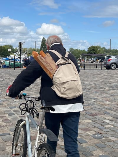 An elderly man with thinning grey hair rides a bicycle with a beige backpack containing two baguettes. He is wearing a black jacket and blue jeans. The bicycle has a black cable lock wrapped around the frame. The man is on a cobblestone street in Honfleur, France, near a harbor with boats and buildings. The sky is blue with white clouds. A silver car is parked in the background.