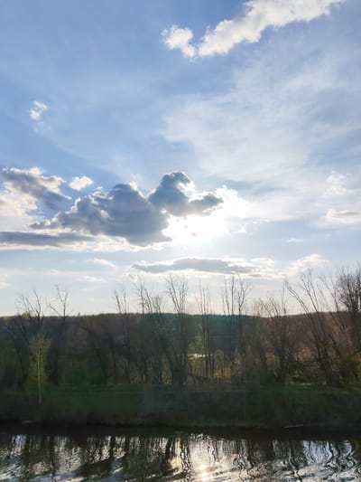 The image depicts a landscape scene near Podosinki, Russia. A body of water, possibly a river or lake, occupies the lower portion of the frame, reflecting sunlight. The shoreline is lined with trees, most of which appear to be bare of leaves. A forested area extends into the background. The sky is predominantly blue with scattered clouds, some of which are obscuring the sun. The sun's rays are visible through the clouds.