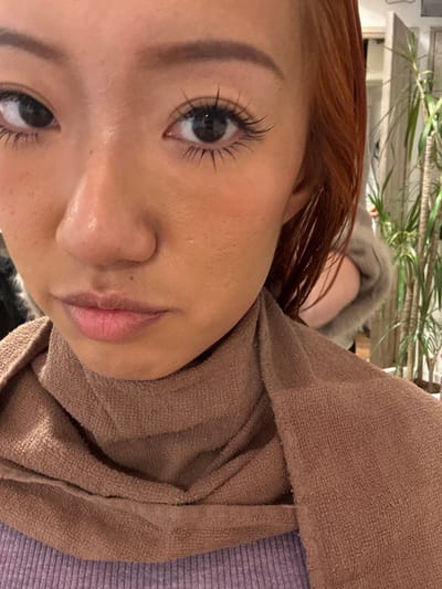 A close-up, head-and-shoulders portrait of a young woman with a medium complexion and reddish-brown hair. She gazes directly forward, featuring defined eyebrows, dark eyes with pronounced, long eyelashes, and subtly blushed cheeks. Her nose and full lips, slightly parted, are visible. A brown terry cloth towel is wrapped snugly around her neck, covering a light purple, ribbed garment. The background is softly out of focus, revealing an indoor environment with several tall green potted plants. A blurred figure, showing a light-colored, possibly fur-trimmed garment on a shoulder and arm, is partially visible behind the subject's right side. The scene is set in an establishment in Toshima, Japan, consistent with a beauty salon or similar personal care venue.