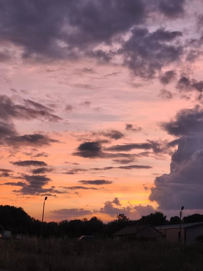 The image captures a twilight sky over Orizari, Bulgaria, characterized by dramatic cloud formations painted in hues of purple, pink, and orange. The sky is the dominant element, with layers of clouds ranging from dark, heavy masses on the right and upper left to lighter, more scattered formations tinged with the colors of sunset across the center and lower portions.

The foreground depicts a silhouetted landscape. A tall, slender lamppost stands prominently near the center, its silhouette sharp against the vibrant sky. To the right of the lamppost, the roofs and walls of several low-rise buildings are visible, also in silhouette. A line of dense trees forms a dark, undulating horizon across the middle ground. The ground in the immediate foreground is covered in dry, sparse vegetation, suggesting a rural or semi-rural setting. Several indistinct vehicles, appearing as dark shapes, are parked in the middle ground. The overall impression is of a quiet, serene evening scene in Orizari.
