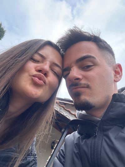 A selfie captured at an upward angle features a man and a woman. The man, positioned at the top of the frame, has dark hair with a shaved undercut and a goatee. The woman, below him, has long brown hair and is puckering her lips in a kissing gesture with her eyes closed. Both appear to be wearing dark-colored jackets. The background reveals a cloudy sky, foliage from a tree, and architectural elements suggestive of a building. The image lacks the identifying elements to confidently suggest it was taken in Straldzha, Bulgaria.