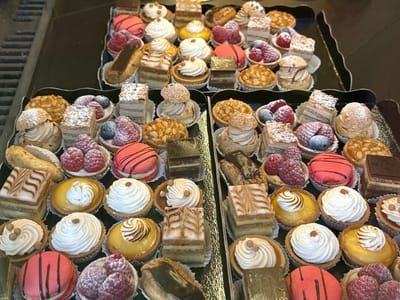 The image displays a close-up, overhead view of a selection of assorted pastries in trays, likely in a bakery or patisserie. The pastries are arranged in multiple rows and columns, with a focus on a variety of small cakes, tarts, eclairs, and macarons. Some pastries are topped with fresh berries, such as raspberries and blueberries, while others feature whipped cream, chocolate decorations, or meringue. The overall presentation suggests a gourmet assortment of individual desserts. The background appears to be a dark, reflective surface, possibly a display case or countertop. The lighting is even, highlighting the intricate details and colors of the pastries. While the image itself does not contain explicit markers of its location, the prompt specifies that it is from Le Lavandou, France.