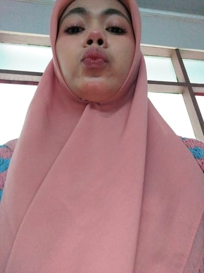 In a close-up, head-and-shoulders shot, a young woman wearing a pink hijab poses for a selfie. Her eyes are closed, and her lips are pursed in a kissing gesture. The background consists of a wall with large, horizontally divided windows. The overall lighting suggests an indoor setting with diffused daylight coming through the windows. There is no discernible indication of the location being Tibojong, Indonesia, in this image.