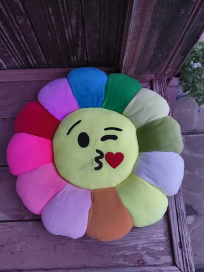 A fluffy, multi-colored flower-shaped pillow with an emoji face is resting on a weathered wooden surface, likely outdoors. The pillow's center is yellow, featuring a winking eye, pursed lips, and a small red heart. Its petals are arranged in a circular pattern and display a spectrum of colors: blue, green, light green, white, pale yellow, orange, light purple, pink, and red. The background consists of dark, vertical wooden planks, suggesting it might be part of a bench or railing in a rustic outdoor setting. The lighting is dim, casting a purplish hue over the scene. The location is specified as Востец Снт, Russia, hinting at a dacha or garden environment.