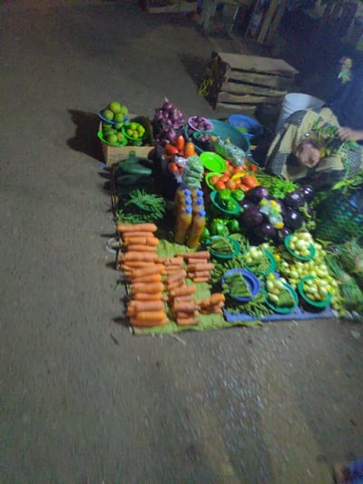 The image shows a street market in Lindi Street, Tanzania, located at coordinates -3.358 latitude and 37.339 longitude. A vendor is selling a variety of produce, including carrots, limes, green beans, eggplant, onions, and other vegetables. The vendor is standing behind the produce, with a blue bucket on the ground next to them. The produce is arranged on the ground on a green mat, with several blue plates scattered around the vegetables. The vendor is wearing a long-sleeved shirt and is interacting with customers. The environment is dark and appears to be at night, with dim lighting.