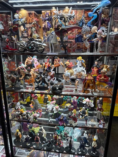 This is a photograph of a retail display cabinet filled with anime figurines. The figurines are arranged on multiple shelves and are of various characters, primarily from the anime series One Piece and Demon Slayer. The display is well-lit, with bright overhead lights illuminating the products. The background consists of shelves filled with more anime merchandise and boxes. The bottom shelf of the display shows some of the packaging for the figurines. The store appears to be an anime merchandise shop. The location is identified as the "Anime Import" store at 139 Avenue de la République, 93420 Villepinte, France.