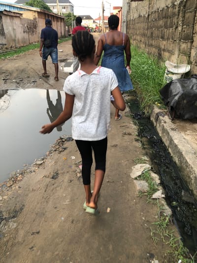A young girl with braided hair, wearing a white patterned shirt and black pants, walks along a pathway adjacent to a water-filled ditch. An older woman in a blue dress walks ahead of her. A man in a blue shirt and shorts can be seen further back. A concrete wall is to the left, and the ditch is on the right. The area is urban with buildings visible in the distance.