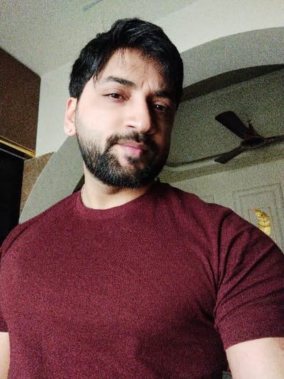 An adult male subject is depicted in a chest-up portrait within an indoor setting in Bidhannagar, India. The individual has dark hair, a full dark beard, and a mustache, with a medium skin tone. He is wearing a maroon short-sleeved, round-neck shirt. A small, silver-colored stud earring is present in his visible left earlobe, and a faint gold-colored chain is discernible around his neck. The subject is looking directly towards the camera. The background reveals a light-colored ceiling with a dark, three-bladed ceiling fan partially visible in the upper right quadrant. An ornate decorative panel, appearing gilded, is visible on the far right wall, and a dark, vertical structural element is present on the left. The lighting suggests an indoor environment.