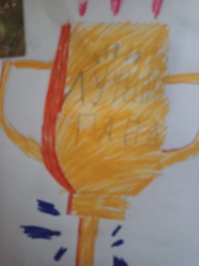 The image is a drawing of a trophy on white paper. The trophy is colored primarily yellow with some red accents on the handles and a red ribbon. There are pink marks at the top of the trophy, possibly representing flames or decorative elements. The trophy has some writing on it, but it is blurred and illegible. There are also some blue scribbles at the bottom of the drawing. The drawing appears to be made by a child. There is no information in the image to suggest a specific location in Novotroitsk, Russia.