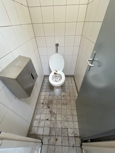 The image shows a small, tiled bathroom stall with a white toilet, a metal paper towel dispenser mounted on the wall, and a gray door with a silver handle. The toilet bowl contains visible feces. The floor is tiled and appears dirty, with scattered debris. The walls are covered in white square tiles with gray grout. The location is identified as a restroom in Bonn, Germany.
