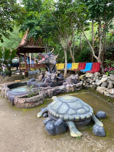 The image depicts a scene in a park or garden located in Kecamatan Manyaran, Indonesia, at coordinates -7.826 latitude and 110.816 longitude. The central focus is a concrete sculpture of a dragon with its mouth open, seemingly roaring, situated in front of a small concrete pond with water. The pond is surrounded by a rocky structure, possibly a representation of a cave or a mountain. To the right of the pond, there is a large concrete turtle sculpture, lying on its back, with its legs visible. Behind the dragon sculpture, a wooden gazebo with a tiled roof stands in the background. There are six colorful chairs aligned in a row in front of the gazebo, suggesting a seating area. The surrounding environment is characterized by lush greenery, including trees and bushes. The ground is covered with gray concrete and scattered rocks, while the overall ambiance suggests a peaceful and playful setting.