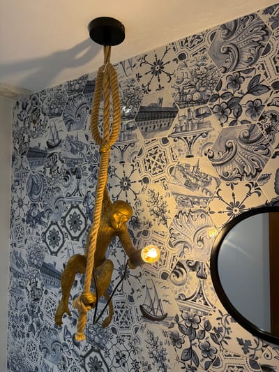 A wall lamp with a monkey figurine and a rope is mounted on a wall covered in hexagonal tiles with a blue and white patterned design. The tiles feature various motifs, including floral patterns, architectural elements, and what appear to be nautical scenes. The monkey figurine is gold-colored and is depicted holding onto a thick rope, which is also part of the lamp's design. A single light bulb is illuminated, casting a warm glow. In the upper right corner, a portion of a circular mirror with a black frame is visible, reflecting the patterned tiles and the light. The overall scene suggests an interior setting, possibly a home or commercial space, with a whimsical and artistic decor. The image appears to be taken indoors, with artificial lighting. There is no visible text in the image. The location context provided is l'Alfàs del Pi, Spain, which might relate to the style of the tiles or the overall aesthetic.