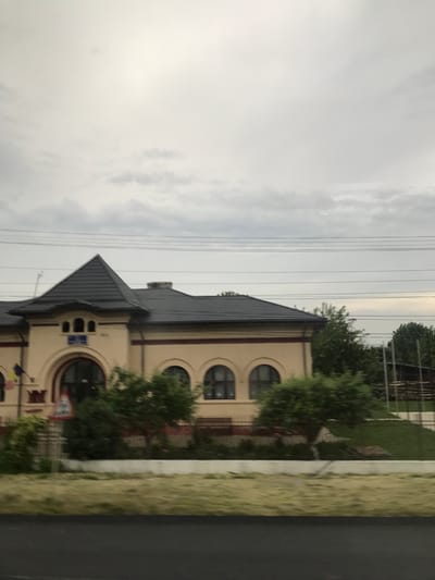 The image depicts a static view of a building and its surroundings in Răcăciuni, Romania, captured from a moving vehicle on a cloudy day. The sky, occupying the upper half of the frame, is overcast with light grey clouds and several dark horizontal utility lines traversing it.

The primary subject is a two-story building constructed of a light beige or pale yellow stucco facade, featuring a prominent dark grey, multi-pitched roof. A distinctive pyramid-shaped roof element is centered above an arched entranceway. Above the entrance, a rectangular blue sign with white lettering is visible, and to its left, a small flag, likely Romanian, hangs from a pole. The building exhibits arched windows with dark frames. A thin, dark red horizontal stripe runs across the upper portion of the facade beneath the roofline. A chimney is visible on the right side of the roof, and a thin antenna-like structure protrudes from the left side.

In the mid-ground, green deciduous trees and bushes are scattered in front of and to the right of the building. A partially obscured red-bordered triangular warning sign is visible in front of the building on the left. Further to the right, beyond the building, additional trees and what appear to be stacked wooden beams or fencing are present on a grassy slope.

The foreground consists of a dark asphalt road surface at the bottom, followed by a strip of pale green to light brown mowed grass, then a low, white concrete wall or curb. The motion blur in the immediate foreground suggests the photograph was taken while in transit. No people or animals are visible, and no specific actions or interactions are depicted.