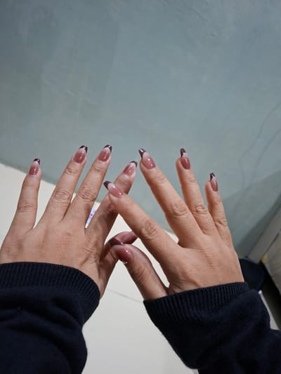Two hands with long, manicured nails are displayed against a grey and white background. The nails are a light nude color with a dark brown French tip design. The hands are positioned with fingers spread, one hand slightly overlapping the other. Both wrists are partially visible, adorned with a dark navy-blue long sleeve sweater. The environment appears to be indoors, with a plain, light-colored surface serving as the backdrop. There is no discernible information to determine the location is in Pie, Indonesia.