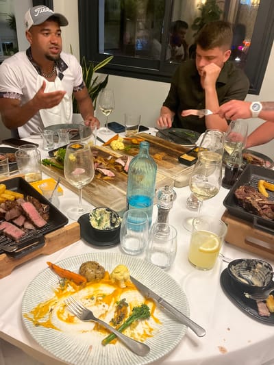The image captures a dinner scene within a building, likely a restaurant, possibly in Urbanització La Cometa, Spain. The table is covered with a white tablecloth and scattered with various dishes, including plates with remnants of vegetables and sauce, a black tray containing cut steak and chips, a wooden platter with roasted potatoes, cauliflower, and other vegetables interspersed with slices of meat, and bowls of salad. Multiple wine glasses, some empty, others containing white wine or lemonade, along with a blue-tinted water bottle, are distributed across the table. Two men are visible: one in a white shirt and baseball cap, gesturing with his hands as if speaking, and another man cutting meat with a fork in his left hand. The room is illuminated by overhead lighting. A window reflects external light.