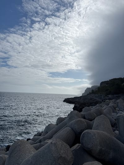 This media file depicts a coastal scene with a focus on a breakwater constructed from large concrete tetrapods. The tetrapods are dark gray, irregularly shaped, and are densely packed, forming a barrier against the water. They appear weathered, with some showing signs of erosion.

The scene is divided into three main areas: the breakwater, the sea, and the sky. The sea is a deep, textured gray, with small waves creating a rough surface. The sky is a mix of dark, moody clouds and patches of brighter, lighter sky. Wispy, white clouds are scattered across the upper portion of the image, suggesting a potentially overcast or changing weather condition. A dark, rocky cliff face covered in sparse vegetation rises behind the tetrapods on the right side of the image.

The perspective is low, looking up towards the cliff and across the water and sky. The orientation of the image is landscape, with the horizon running vertically. There is no visible text in the image. The overall impression is one of a rugged, natural coastal environment, possibly a harbor or a stretch of coastline protected by the breakwater. The lighting suggests it could be daytime, but the cloudy conditions create a somewhat somber atmosphere.