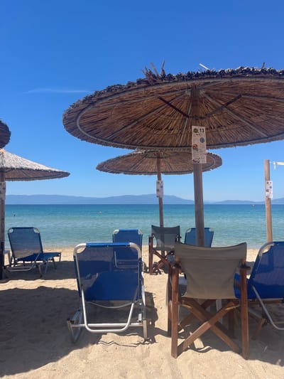 The image depicts a serene beach scene at Ofrynio Beach, Greece, under a clear blue sky. In the foreground, several beach chairs and thatched umbrellas are arranged on the sandy shore, indicating a relaxing atmosphere. The chairs are made of blue fabric and metal frames, with one wooden director's chair in the center. The umbrellas, constructed from natural materials, provide shade. Beyond the chairs, the turquoise waters of the sea stretch towards the horizon, where distant mountains are visible. The water is calm, with a few subtle ripples. There are two individuals visible in the water, one appears to be swimming, while another is further out. The scene is characteristic of a well-maintained beach resort, likely during a sunny day. Signs are attached to the umbrella poles, suggesting services or information for beachgoers. The overall impression is one of tranquility and leisure.