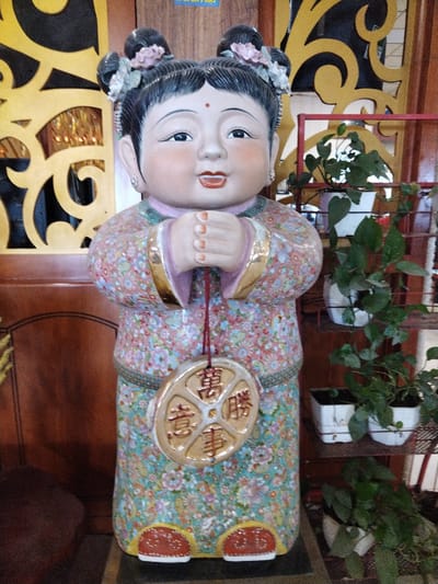 A ceramic figurine of a young Chinese girl stands in an indoor setting, likely in a business in Chu Văn An Ward, Vietnam. The girl is wearing a floral-patterned robe with gold trim and holding a round, golden pendant inscribed with Chinese characters. She has black hair styled in buns adorned with pink flowers, a fair complexion, and a red dot on her forehead. A dark wooden door with intricate gold accents is visible behind her on the left. To her right, a metal shelving unit holds small potted green plants in white square pots. The lighting is soft and ambient.