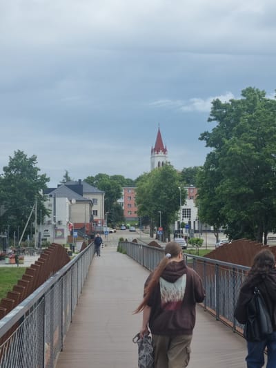 The image captures a scene on a pedestrian bridge in Kuršėnai, Lithuania, on an overcast day. Two individuals are walking away from the camera on the bridge, which has metal railings and a wooden walkway. The person in the foreground has long, light brown hair tied in a ponytail and is wearing a brown hooded sweatshirt with a graphic design of reeds on the back, and khaki cargo pants. They are carrying a patterned tote bag. The second person is wearing a dark hooded sweatshirt and a black backpack. In the background, buildings and trees line a street. A distinct white tower with a red steeple, likely a church spire, is visible above the trees. The overall environment suggests a temperate climate, with lush green foliage.