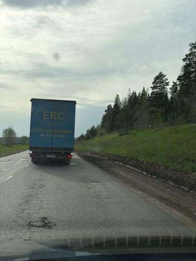 The image depicts a blue delivery truck on a road in Staryi Chul'tem, Russia. The truck has yellow lettering on its side that reads "ERC ENGLJAHRINGER Road Cargo." The sky is cloudy and there are trees lining the side of the road. The road surface appears wet. The image was likely taken from a vehicle moving behind the truck.