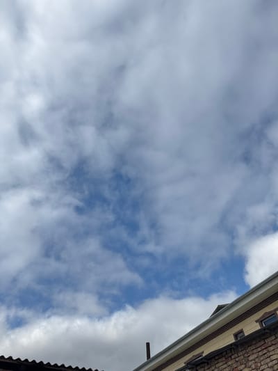 The image showcases a view looking up at the sky, partially obscured by soft, billowing white clouds. Patches of blue sky are visible in between the clouds. A fragment of a building is visible on the upper and lower right corner of the frame. The building features a beige façade, brickwork, and several windows.