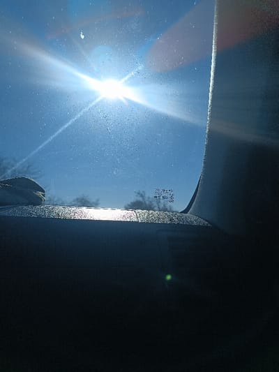 The image presents a view from inside a vehicle located in Gamarjveba, Georgia, looking out through a sun-dusted windshield. A bright, central sun flare dominates the upper-left quadrant of a clear blue sky, casting strong light and revealing numerous speckles on the glass. The vehicle's dark dashboard occupies the lower portion of the frame, with a band of sun glare reflecting across its top surface. A light-colored object, possibly a folded towel or cloth, rests on the dashboard near the left edge. The dark, vertical A-pillar of the vehicle is visible on the right side. Faint outlines of distant, bare trees or shrubbery are discernible through the windshield in the middle ground. Illegible technical markings are etched into the windshield glass in the mid-right. A green optical lens flare is visible on the dark dashboard in the lower center. No people are visible.