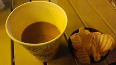 A still life photograph features a yellow paper cup filled with a light brown beverage, likely coffee or tea, placed to the left of a small black bowl containing ridged potato chips. The cup has a subtle decorative pattern visible. Both items rest on a yellow surface with a slatted texture, suggesting an outdoor table. The lighting is dim and warm, casting soft shadows. No individuals are present, and no actions are depicted. The scene is devoid of any contextual clues indicating a specific location within Baubau, Indonesia.