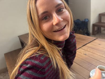 A close-up, frontal image captures a female individual indoors in Porto Salvo, Portugal. The subject, a woman with long blonde hair and a fair complexion, occupies the majority of the frame, facing the camera with a wide smile, displaying her teeth. She is wearing a high-neck sweater patterned with alternating burgundy and gray chevron stripes. Her right hand is partially visible in the bottom right corner of the frame. The immediate foreground features a light-colored wooden table with a distinct plank pattern. On the table, to the right, a white espresso cup on a matching saucer contains a small amount of dark liquid. Adjacent to the cup is a small white packet. The background consists of a light-colored wall to the left and a darker area to the right, where dark wooden furniture, possibly benches or table bases, is discernible. An indistinct dark object is present in the distant background.