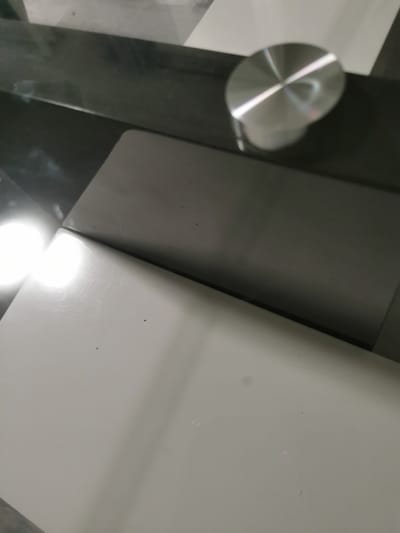 The image depicts a close-up view of a modern desk or table surface in what appears to be an indoor setting. The surface is divided into two distinct sections: a darker, possibly glass or polished wood section on the upper left, and a lighter, matte surface on the lower right.
On the darker section, a circular metallic object with concentric rings and a bright reflection is visible. This object is likely a decorative element, a foot of furniture, or a component of a desk lamp. The lighter section of the surface shows some minor imperfections, including a few small dark specks and faint scratches. A bright, circular light source is reflecting intensely on the left side of this lighter surface.
The overall impression is one of a clean, contemporary workspace or furniture item. There is no indication of the specific location within the city of Bourg-lès-Valence, France, in the provided image. The scene is static and shows no human presence or activity.