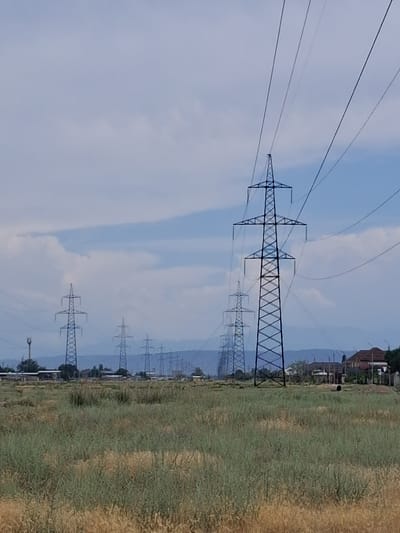 An outdoor, daytime, medium-angle shot captures a row of large electricity pylons stretching across a vast, dry, grassy field under a cloudy sky. The pylons are metallic, lattice-style structures with thick cables stretching between them and disappearing into the distance. In the background, a few buildings, including houses with red tile roofs, are visible, along with a line of distant mountains. The overall scene appears to be on the outskirts of a city, with the pylons indicating power lines that likely serve the area. This image is consistent with the landscape found near Taraz, Kazakhstan, an area characterized by open fields and agricultural surroundings, often featuring such infrastructure to support its population and industry.