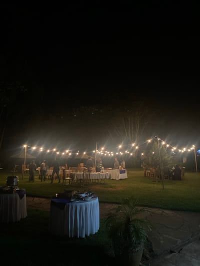 The image captures an outdoor evening gathering, likely a dinner or party, taking place on a grassy lawn illuminated by a long string of warm fairy lights. The scene is set at night, with artificial lighting being the primary source of illumination.
In the foreground and extending into the midground, several long tables are set up, covered with white tablecloths, some with blue accents underneath. These tables appear to be serving stations, with various dishes and serving equipment, including chafing dishes, visible on them. Scattered around the tables are numerous chairs, and several people are present, either standing, serving themselves, or seated, though their faces and specific activities are indistinct due to the distance and low light.
The grassy area is well-maintained, and there are trees and foliage visible in the background and to the right, including a prominent palm-like plant in a large pot or planter on the right side. The ground to the far right appears to be paved or tiled. The string lights, stretching across the frame, cast a festive glow over the event, creating a warm and inviting atmosphere. No discernible text is visible in the image. The setting suggests a celebratory or social function in a garden or backyard environment, potentially in Riara Ridge, Kenya as indicated by the context.