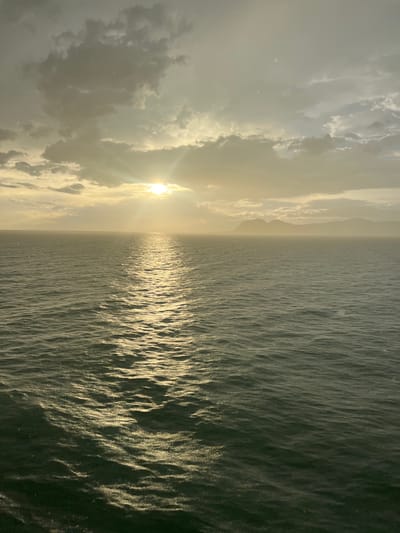 The image depicts a serene seascape at sunset, likely near Don Sak, Thailand, given the context. The sun, partially obscured by clouds, casts a warm, golden light across the scene, creating a shimmering reflection on the gently rippling water. In the distance, a hazy silhouette of mountains or landmasses can be seen against the twilight sky. The sky itself is a mix of pale yellow and gray clouds, with visible rays of sunlight breaking through. The overall atmosphere is tranquil and evokes a sense of peace. There are no people or man-made objects prominently featured in the image, focusing solely on the natural elements of the ocean, sky, and distant land. The water appears dark green, with the sun's reflection creating a bright, broken path across its surface.