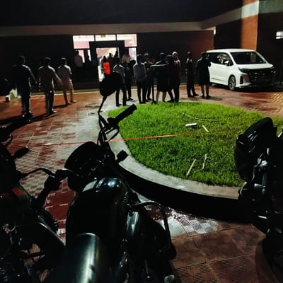 The photo was taken at night in Lalmohan, Bangladesh. A group of people stand near a building entrance, while others walk towards it. A white minivan is parked on the right, adjacent to a small patch of grass. Two motorcycles are in the foreground, partially obscuring the view. The ground is paved with a textured, brick-like pattern.