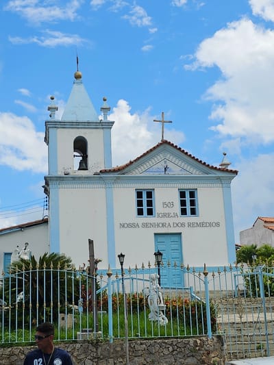 This image depicts the Nossa Senhora dos Remédios church located in Arraial do Cabo, Brazil, viewed under a bright blue sky with scattered white clouds. The church is a white structure accented with light blue trim and features a prominent bell tower on the left with a conical light blue roof topped by a golden finial, inside which a bell is visible. The main facade displays two rectangular windows with light blue frames, a central light blue double door, and a triangular pediment above, containing a tiled mosaic of a standing figure. A wooden cross surmounts the pediment. Inscriptions on the facade read "1503", "IGREJA", and "NOSSA SENHORA DOS REMÉDIOS". The roof is composed of reddish-brown tiles.
In the foreground, a light blue metal fence with golden decorative spikes separates the viewer from the church property. Behind the fence, a garden area features various green plants, bushes, and flowering flora with red and yellow blooms. Two black ornamental street lamps are situated within the garden. Two white statues are visible within the fenced area: one on the left depicting an angelic figure holding an object, and another on the right showing a robed figure. Stone steps are partially visible leading upwards on the right side of the frame. Power lines are present on the far left. The immediate foreground consists of a stone wall.
In the lower left corner, partially visible, is a male subject wearing sunglasses, a dark shirt with the number "02" printed on it, and a necklace. He is oriented towards the right.