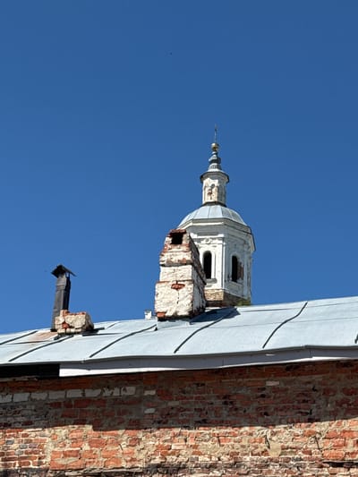 The image shows the top of a brick building with a metal roof, against a clear blue sky. On the roof, there are two chimneys. To the right of the chimneys is a white dome with a spire, which appears to be part of a church or a bell tower. This dome has some decorative architectural elements and arched windows. The brickwork of the building is weathered, showing signs of age. The scene is likely part of the historic architectural landscape of Yelets, Russia, which is known for its numerous churches and historical buildings.